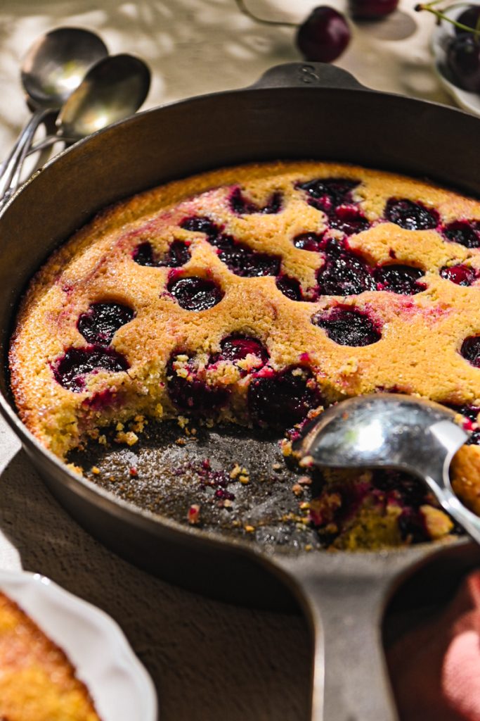 Serving spoon taking some cherry spoon cake from a cast iron skillet.